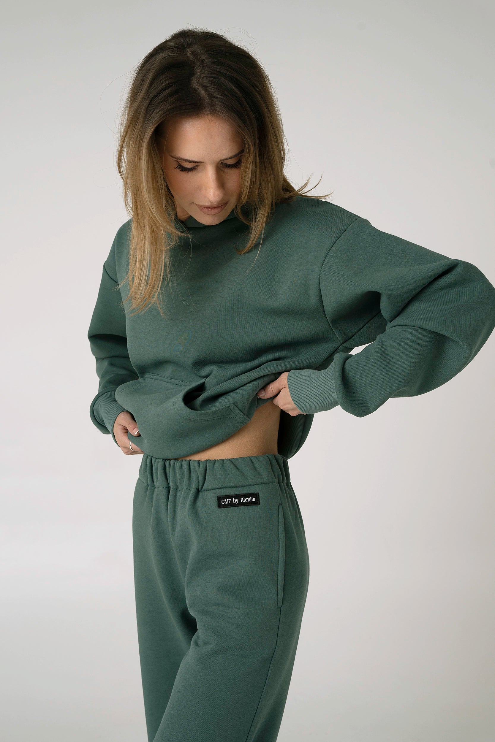 Žalios spalvos laisvalaikio treningų komplektas moterims – džemperis be užtrauktuko ir kelnės su kišenėmis, modelis rodo detalų audinio vaizdą.