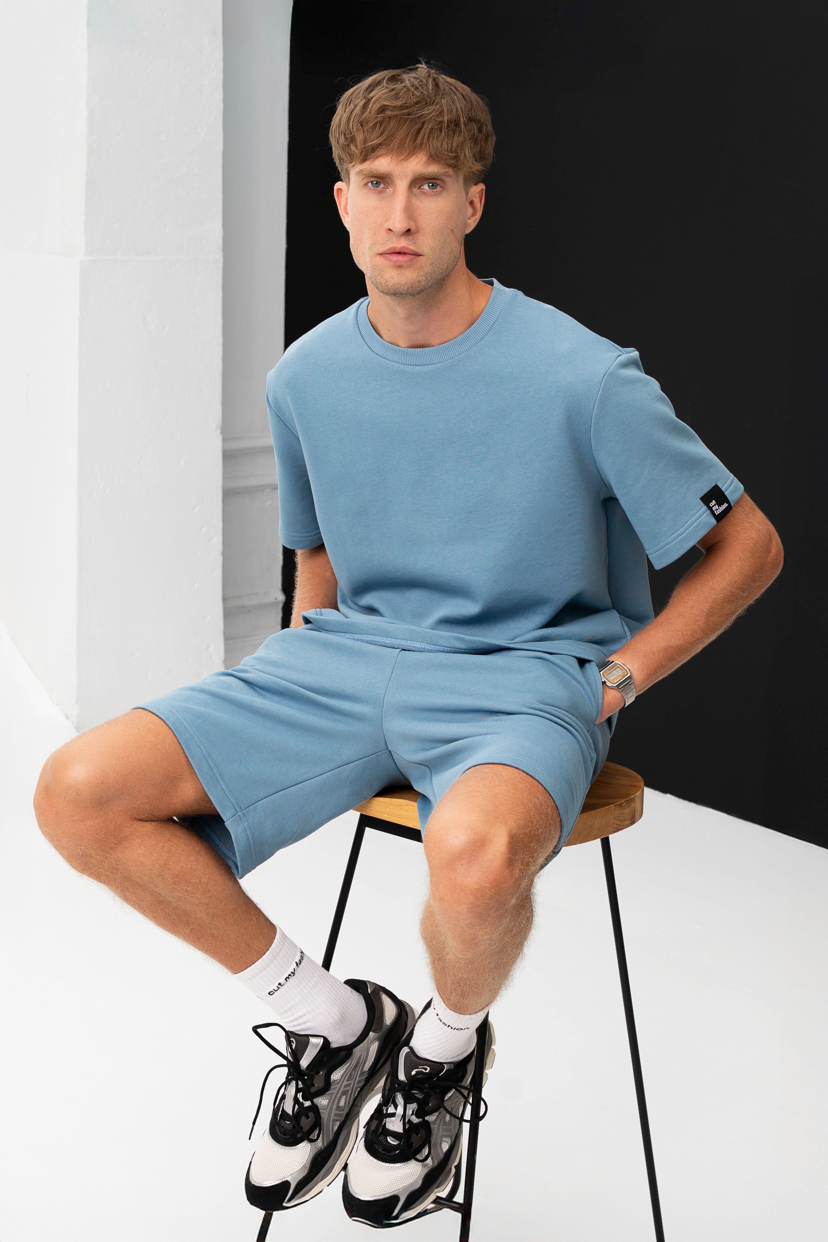 Modelis sėdi ant kėdės dėvėdamas šviesiai mėlyną laisvalaikio kostiumą su šortais, rankos kišenėse