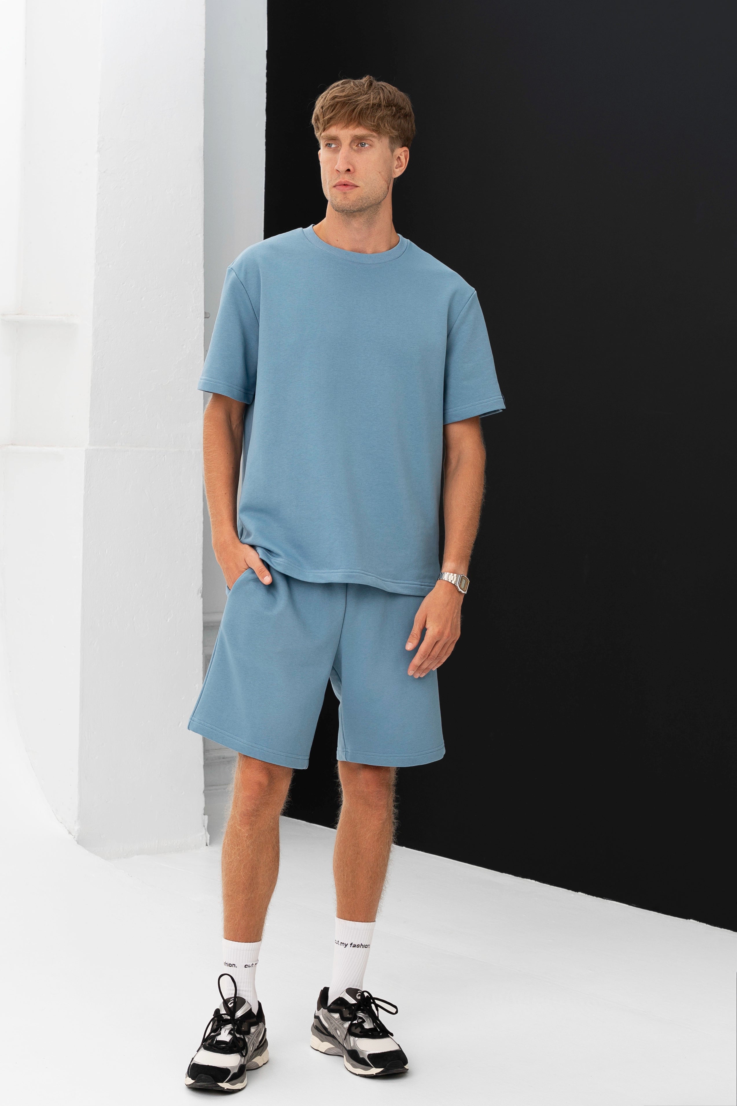 Modelis pozuoja su šviesiai mėlynu „Aras“ treningų komplektu – marškinėliais ir šortais, rankos kišenėse