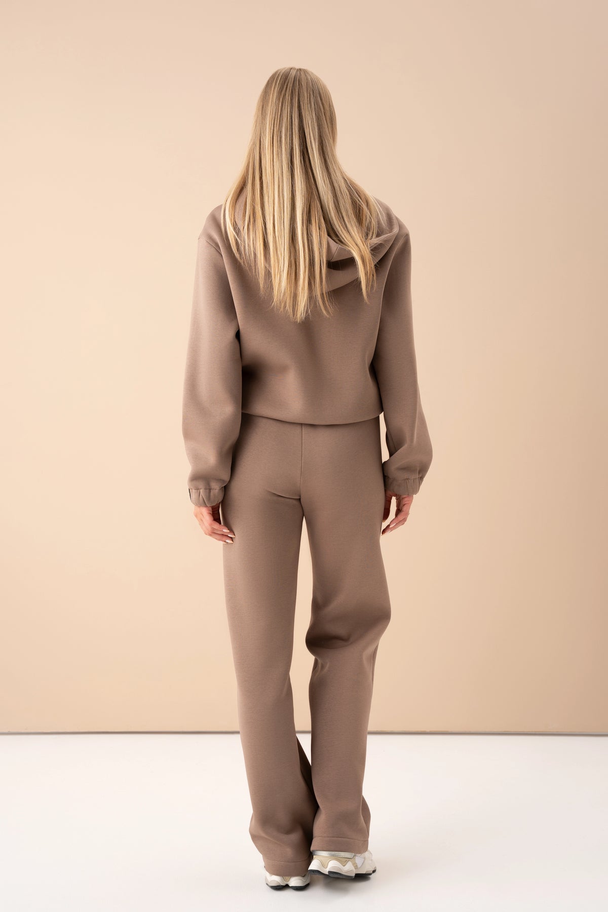 woman wearing a latte-colored cutmyfashion tracksuit, turned with her back to the camera, posing against a light studio background