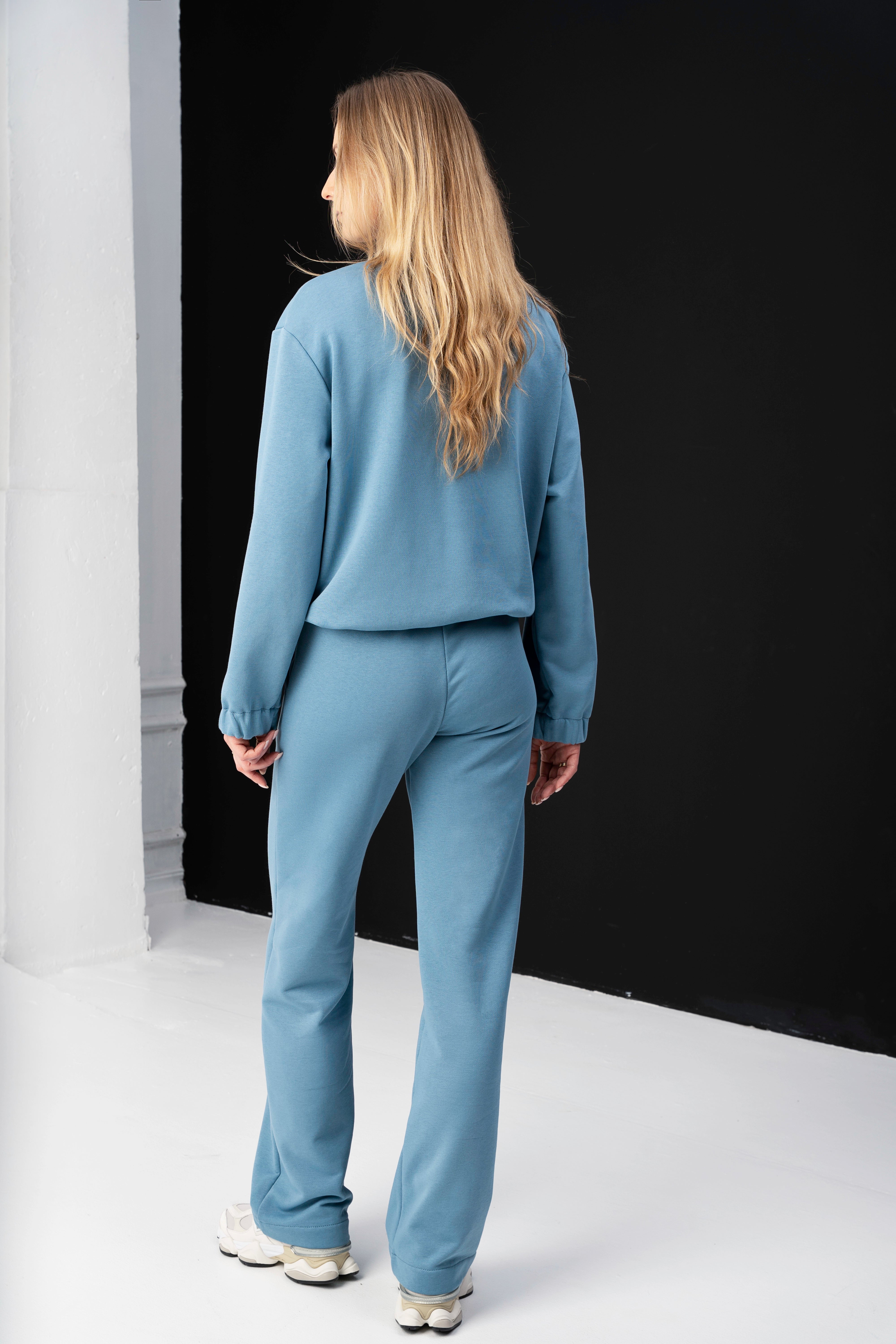 modelis apsirengusi oversized mėlynu laisvalaikio kostiumu, stovi priejuodos sienos, pozuoja nugarą