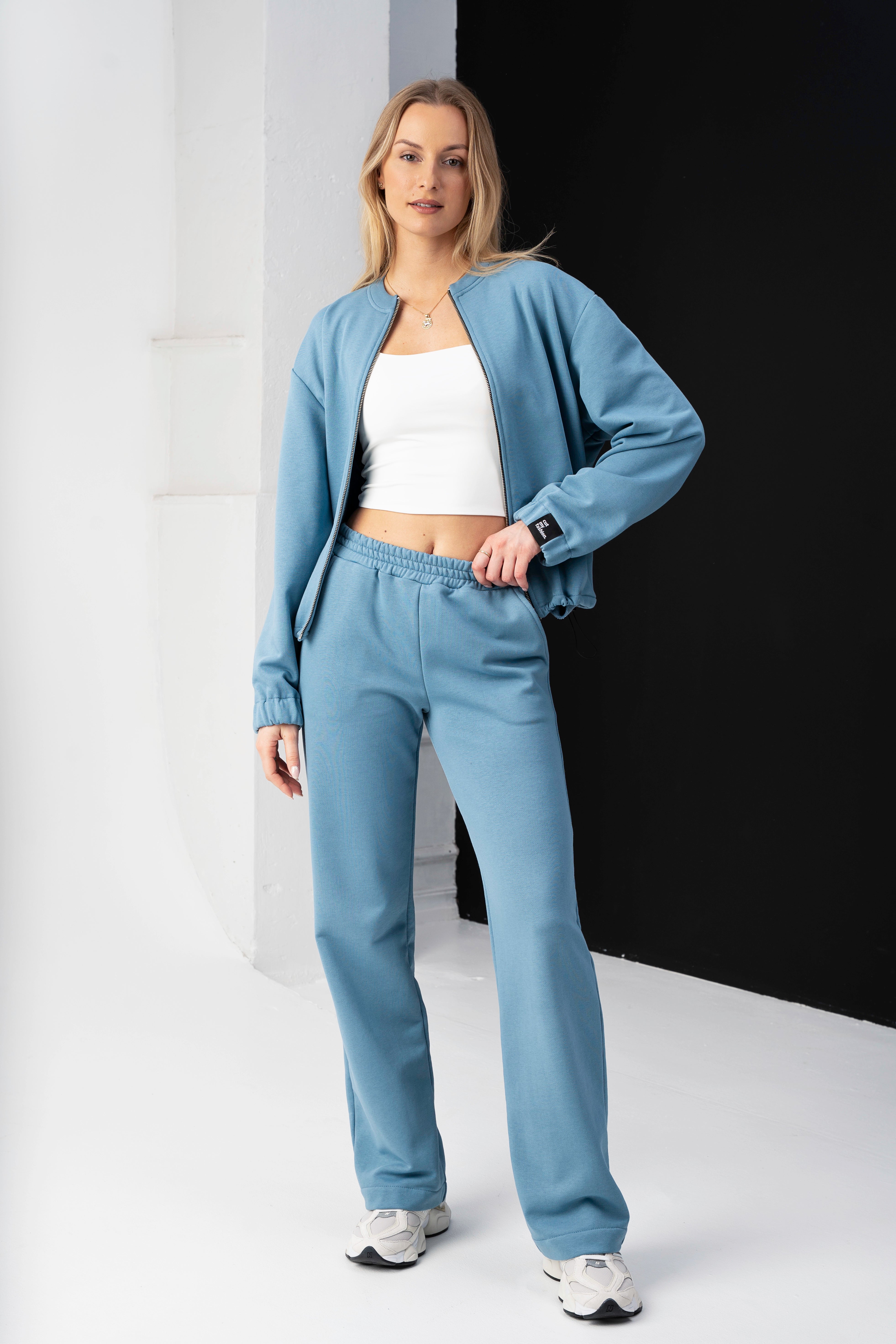 moteris vilki mėlynus treningus, ranka prilaiko kelnių juosmenį, matomas baltas topas