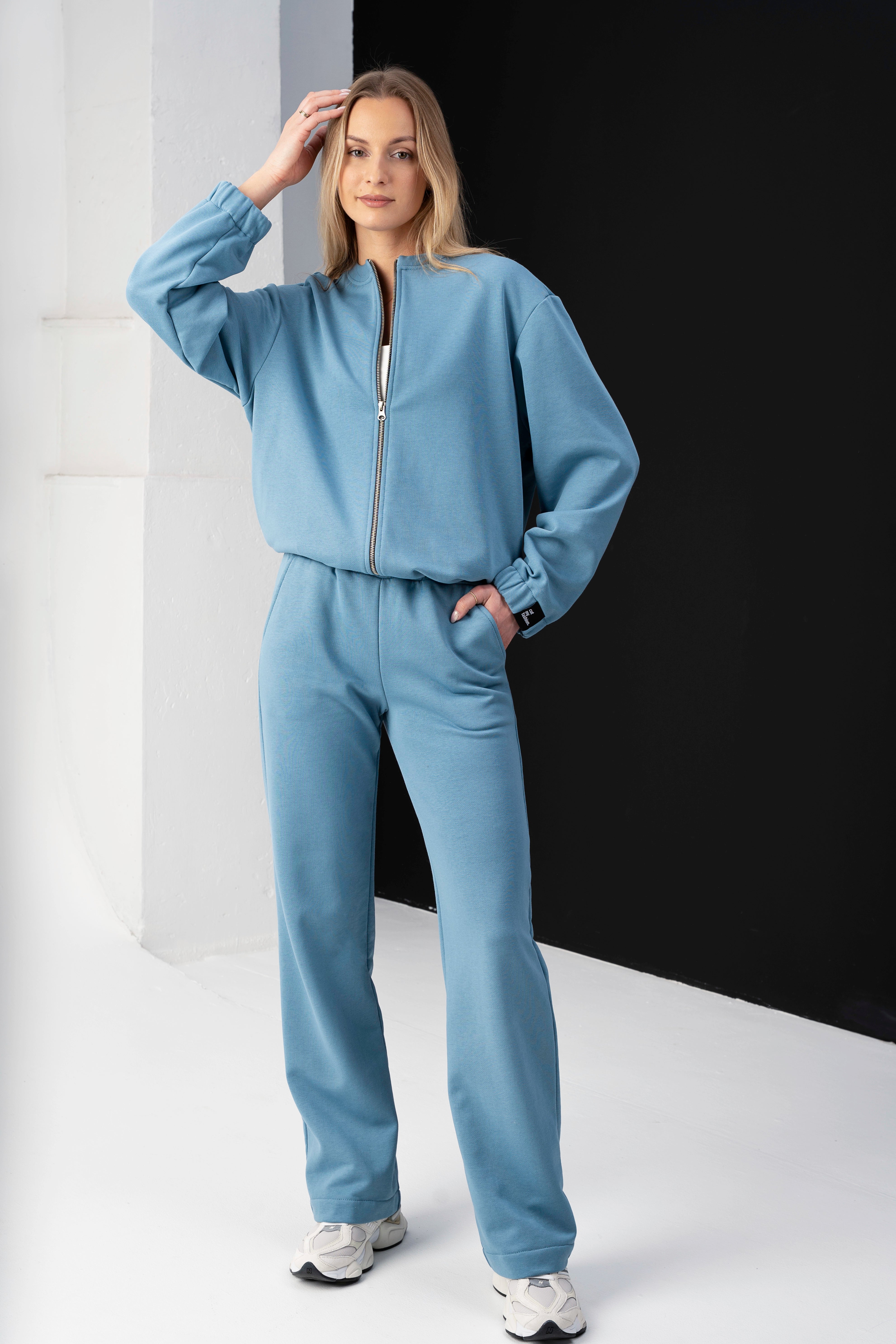 modelis dėvi laisvalaikio kostiumą su šviesiai mėlynu džemperiu ir kelnėmis, ranką laiko ant galvos, stovi juodame fone