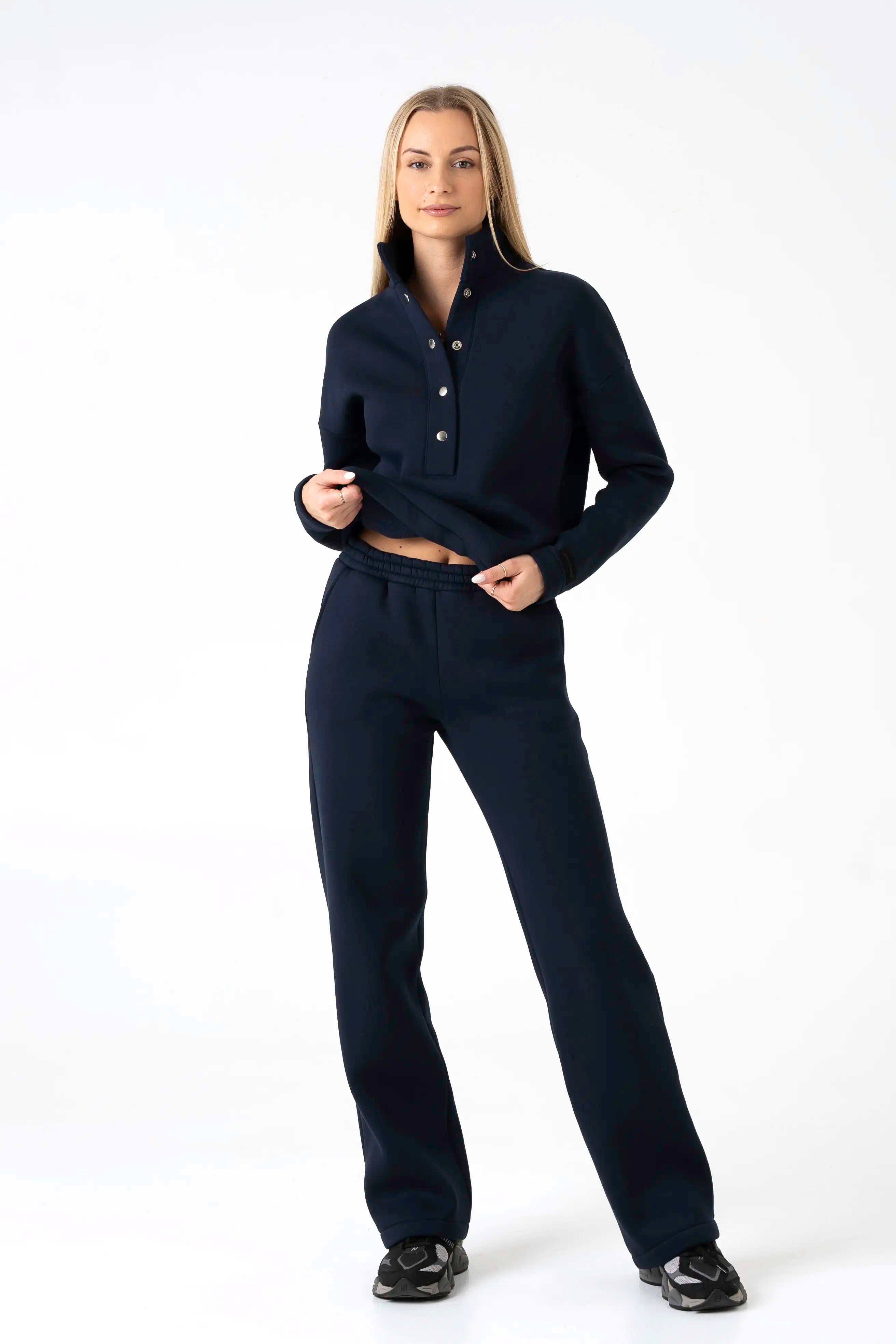 Modelis stovintis ir šiek tiek prilaikantis mėlynos spalvos cutmyfashion laisvalaikio treningų viršų su sagomis, demonstruojantis komfortišką stilių