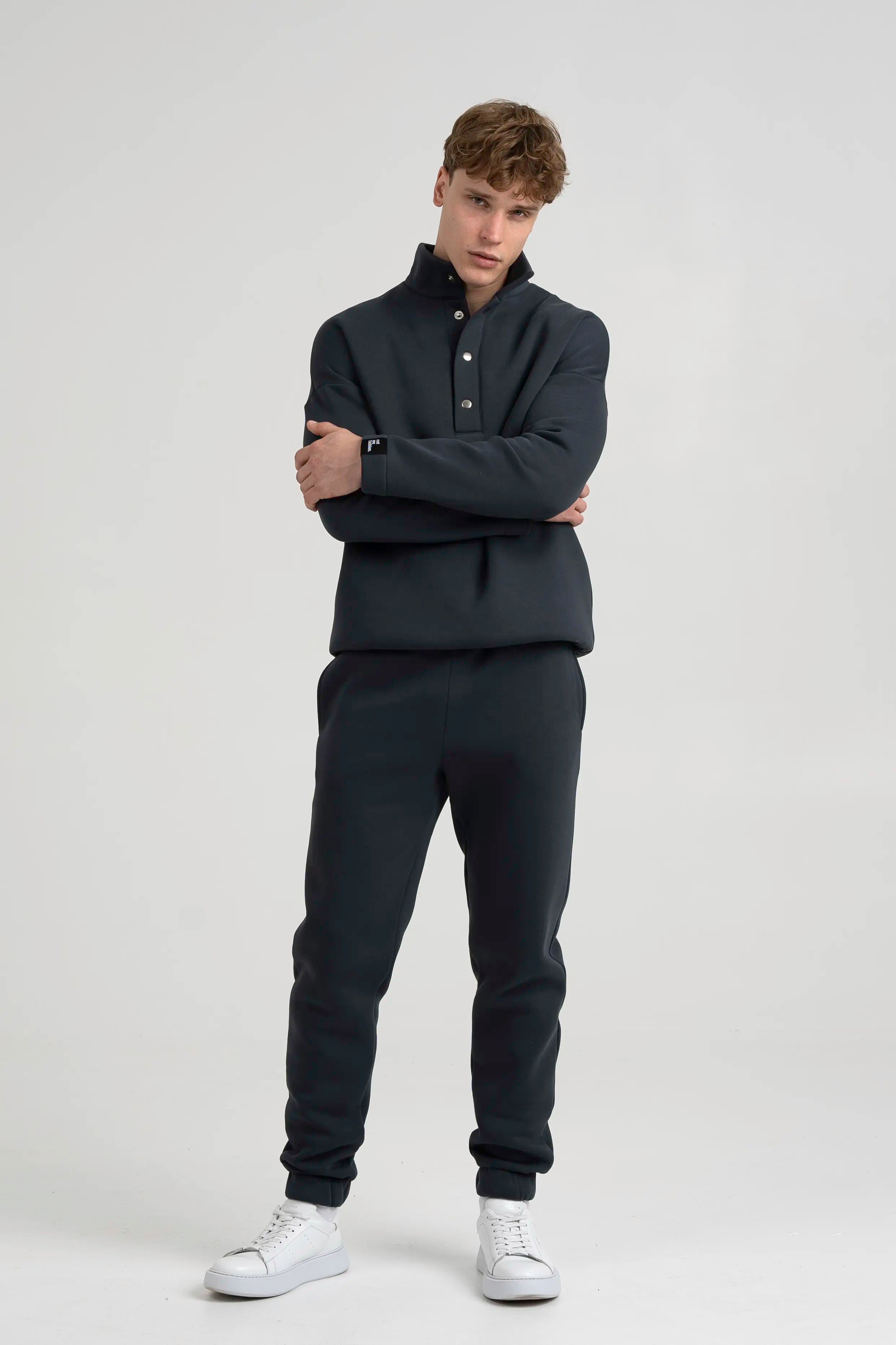 Vyras modelis su tamsiai pilkais laisvalaikio treningais, su sagomis ties apykakle ir šoninėmis kišenėmis, susikabinęs rankas priekyje