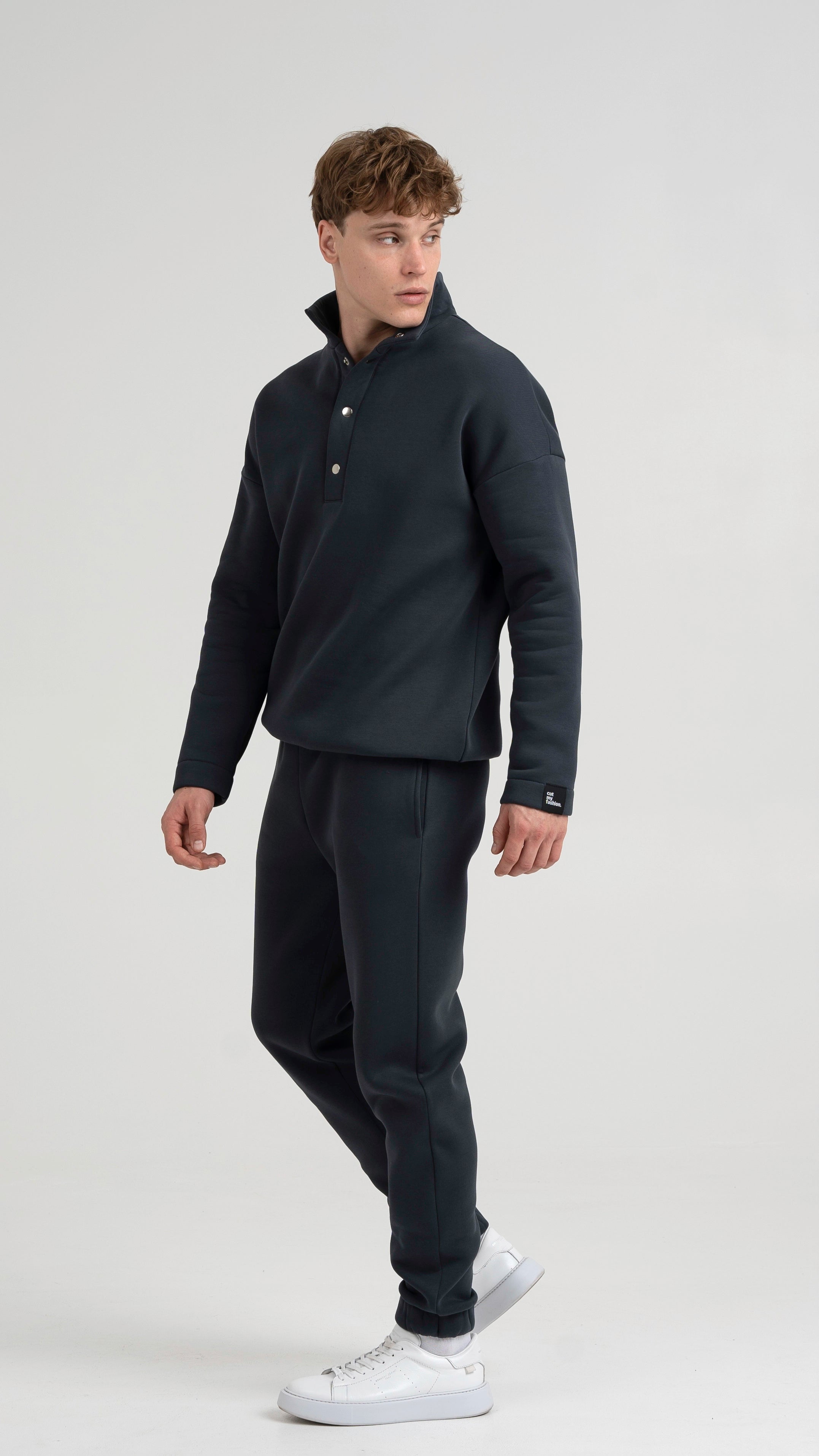 Vyras iš šono profilio su tamsiai pilkais treningais, pabrėžiamas laisvas kirpimas ir susegamos kelnės su baltais sportbačiais