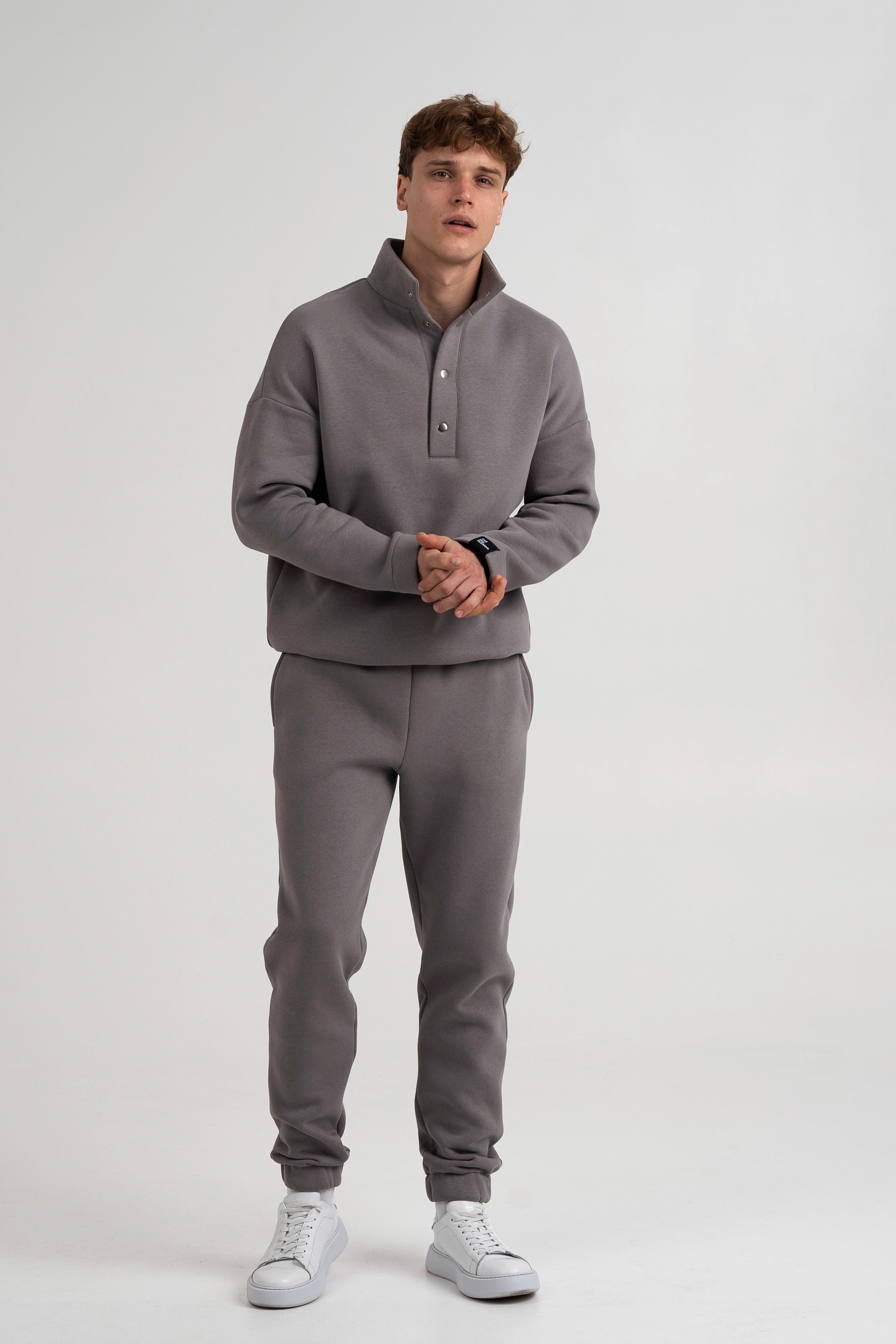 Modelis dėvi cutmyfashion pilkos spalvos vyriškus laisvalaikio treningus su tiesiomis kelnėmis, demonstruojant patogų ir minimalistinį stilių