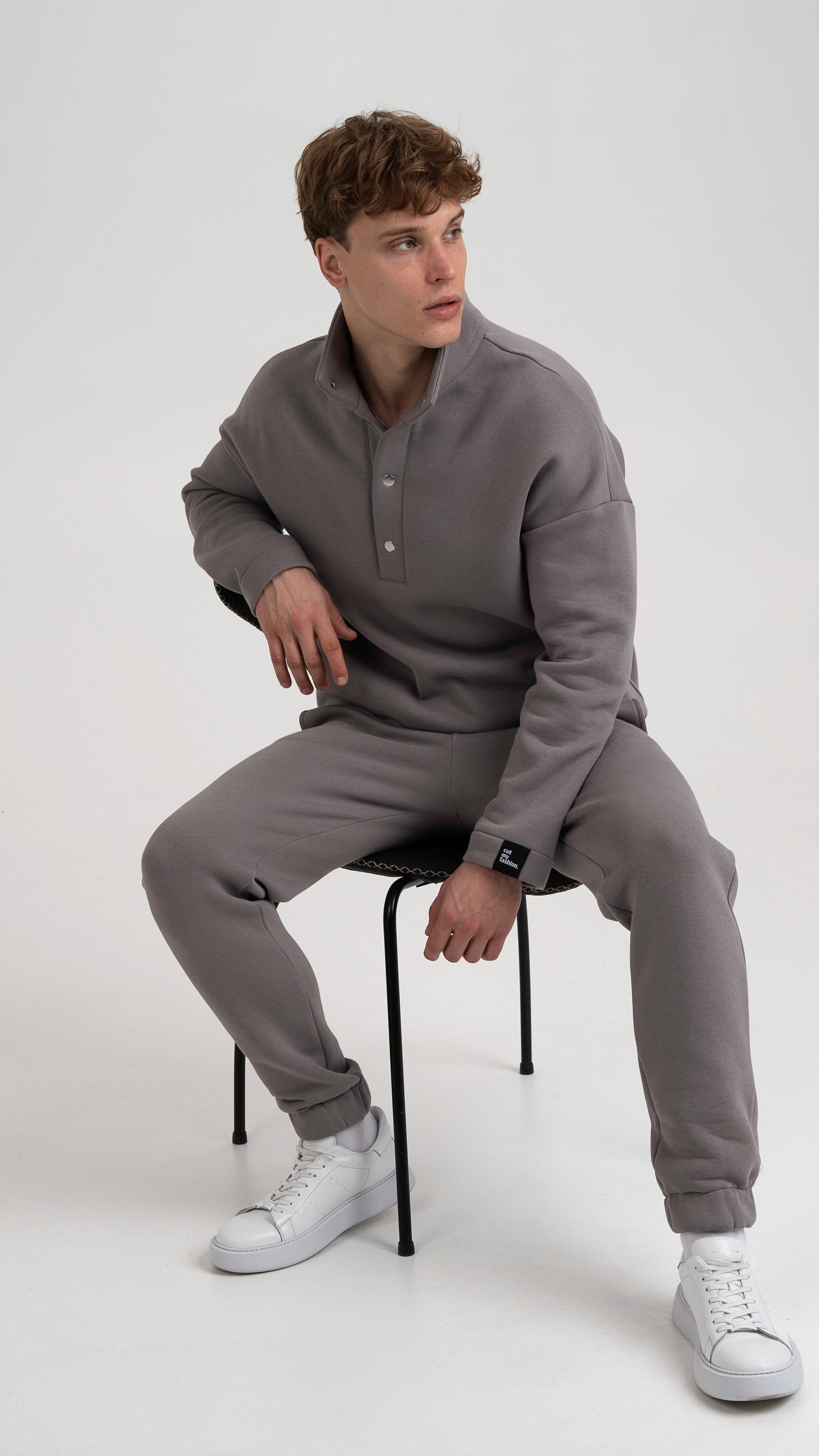 Modelis sėdi ant kėdės, dėvint Cutmyfashion pilkos spalvos vyriškus laisvalaikio treningus, demonstruojant stilingą patogumą