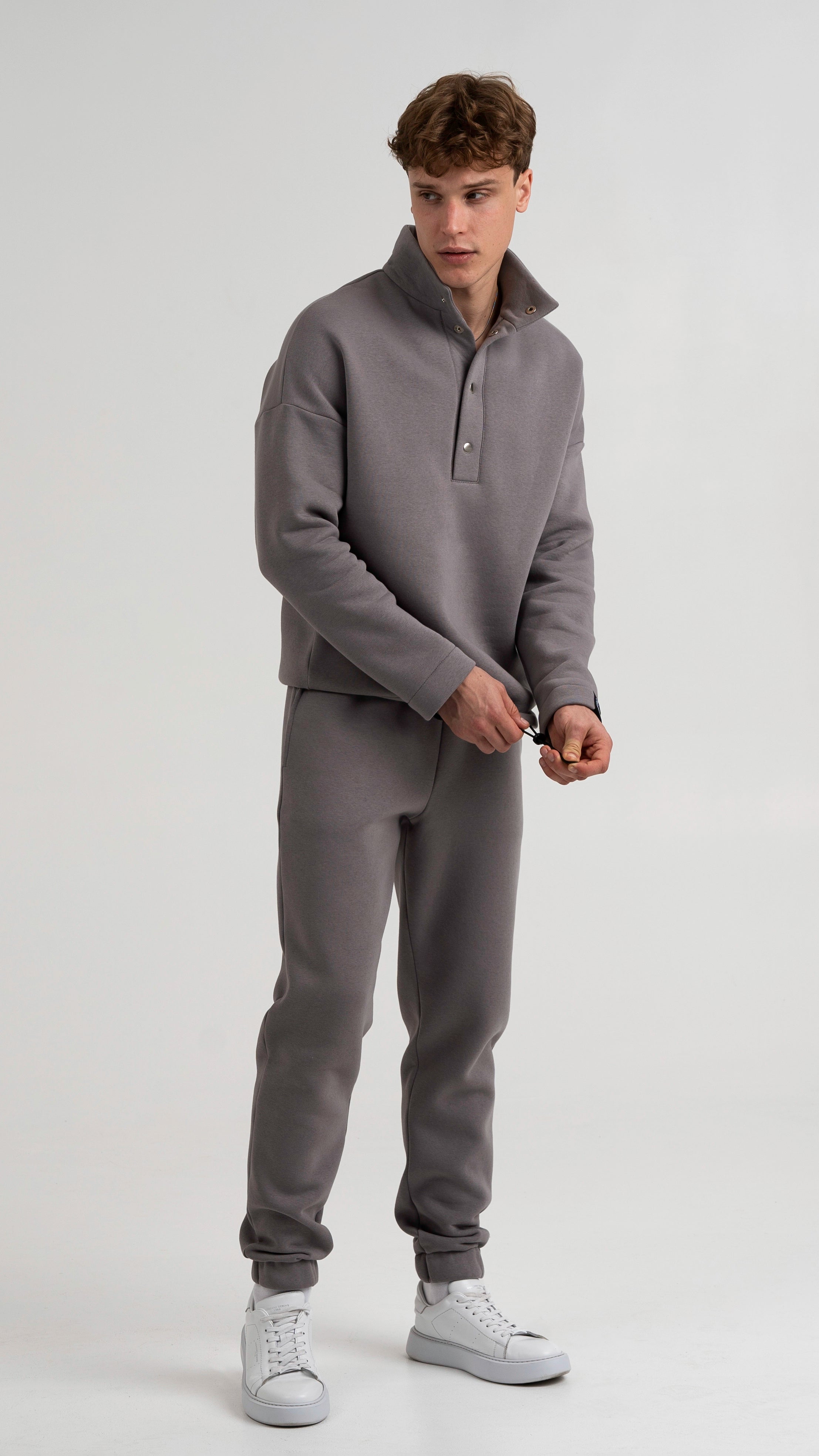 Modelis dėvi Cutmyfashion pilkos spalvos vyriškus laisvalaikio treningus su sagomis ir surauktomis kelnėmis, stovėdamas ir laikydamas raištelius