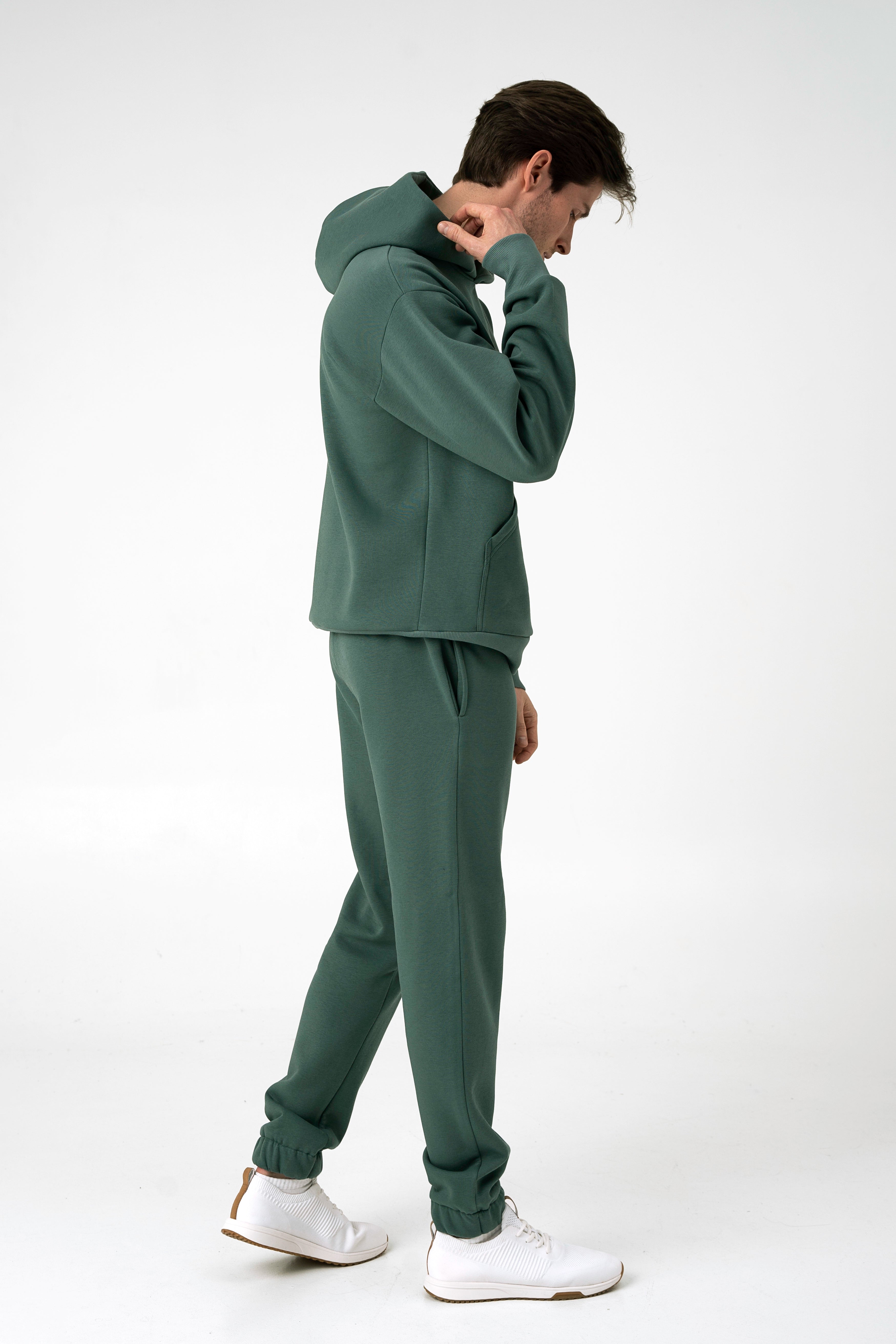 Vyras stovi pasisukęs šonu ir ranka laiko už džemperio kapišono baltame fone ir dėvi CMF By Kamilės laisvalaikio treningus CMF184