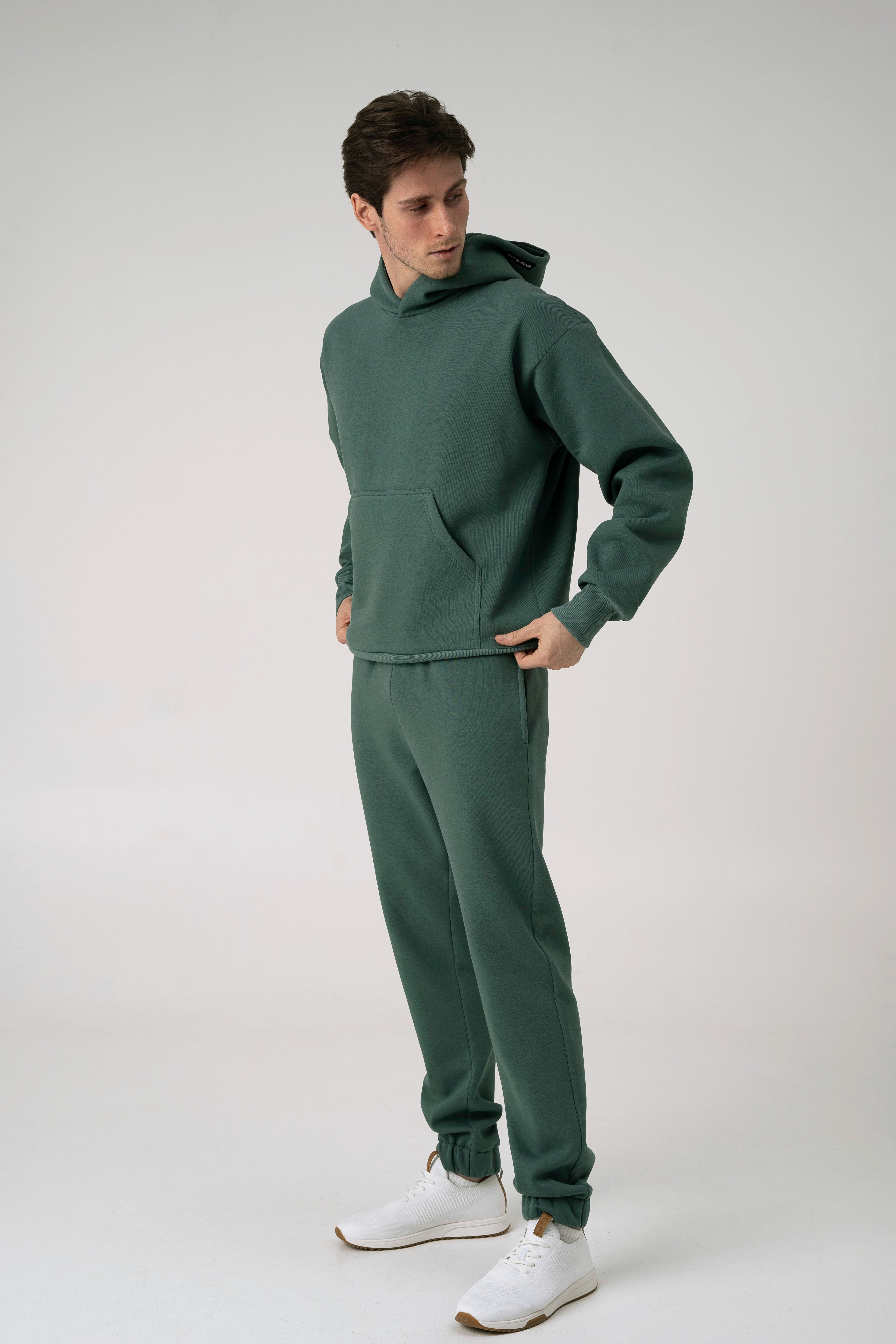 Vyras stovi pasisukęs šonu baltame fone ir rankas laiko už džemperio apačios ir dėvi CMF By Kamilės laisvalaikio treningus CMF184