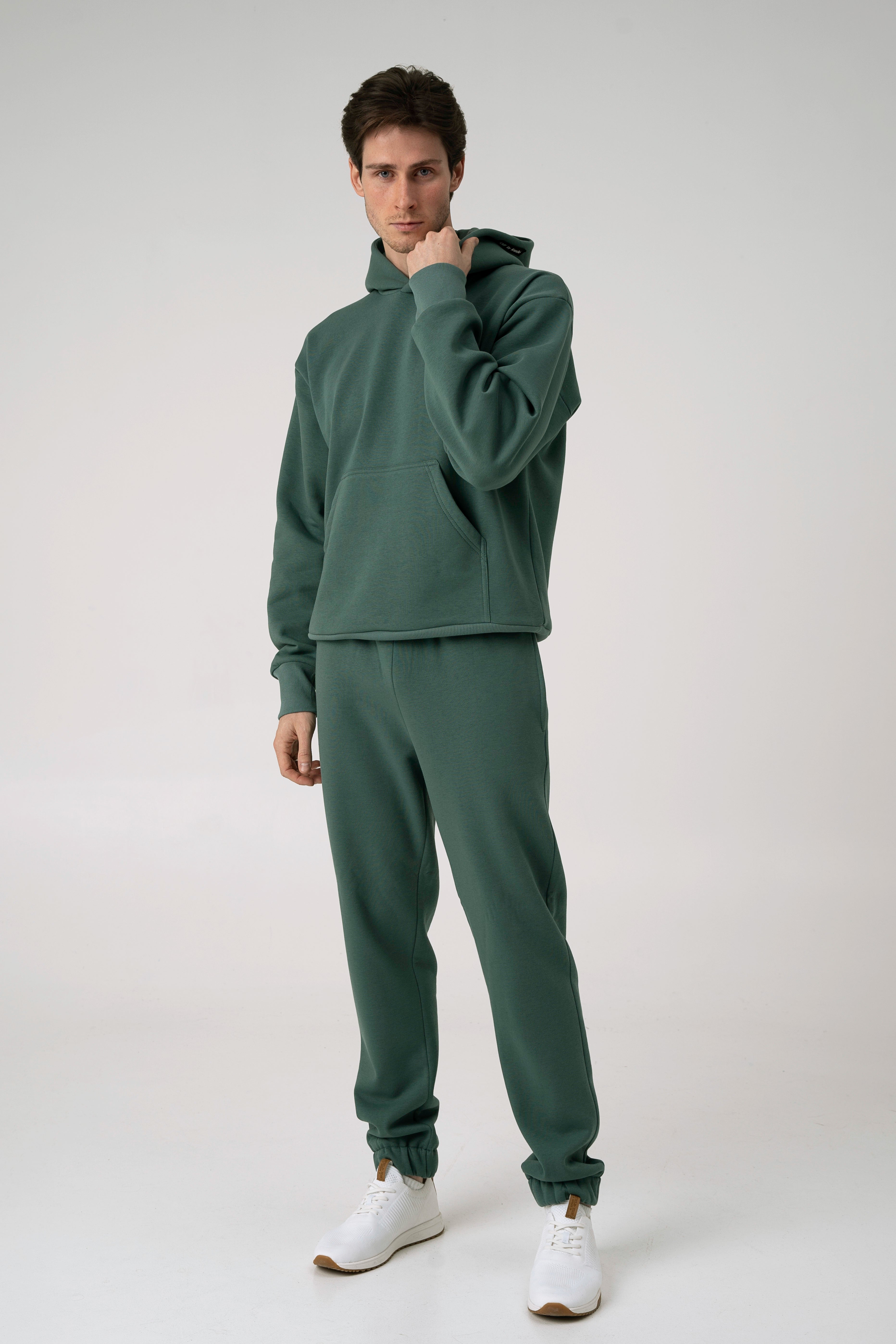 Vyras stovi ir ranka laiko džemperio kaklą baltame fone ir dėvi CMF By Kamilės laisvalaikio treningus CMF184