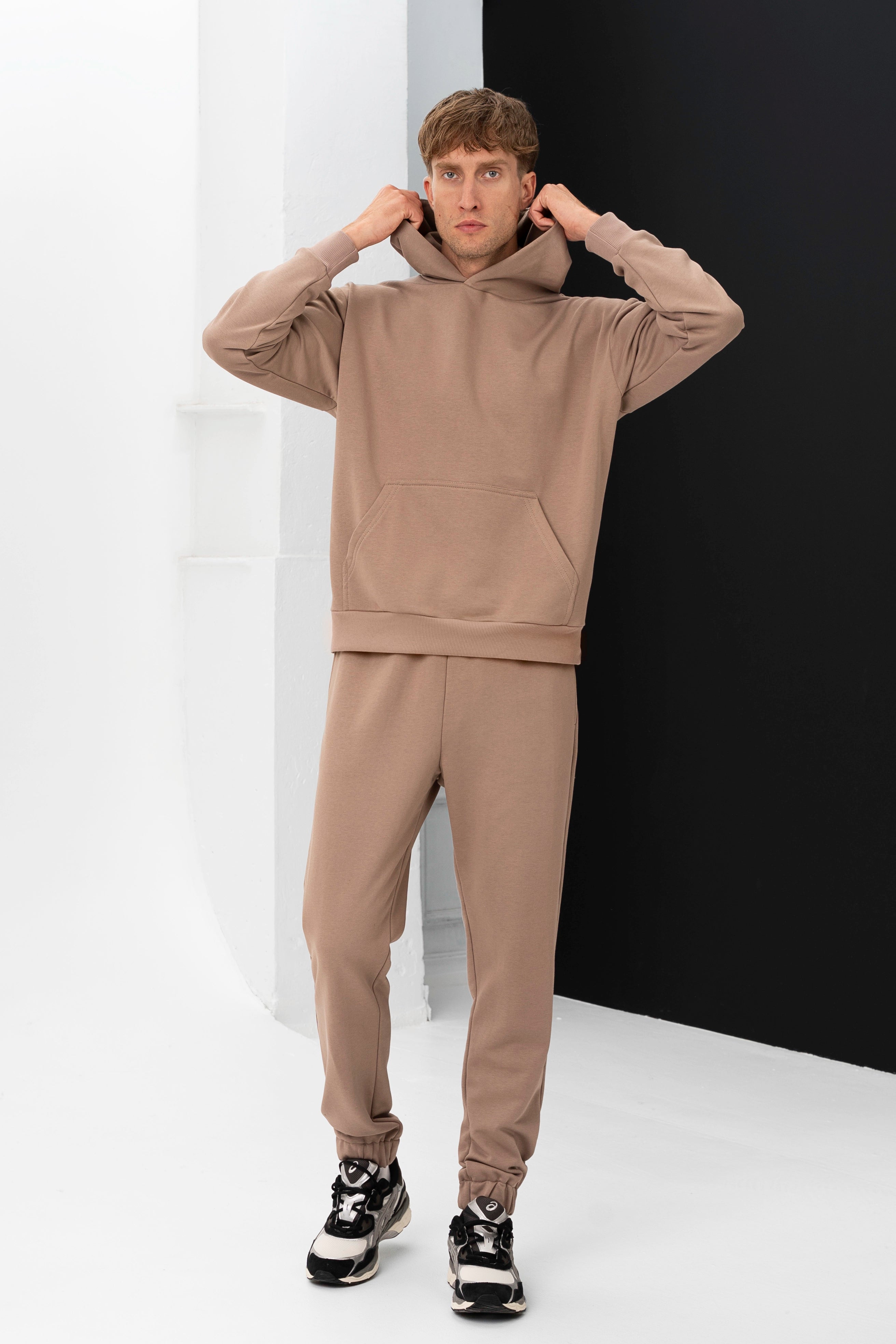 Modelis laiko rankomis už džemperio kapišono, vilkėdamas smėlio spalvos vyriškus treningus, stovėdamas prieš kontrastingą foną