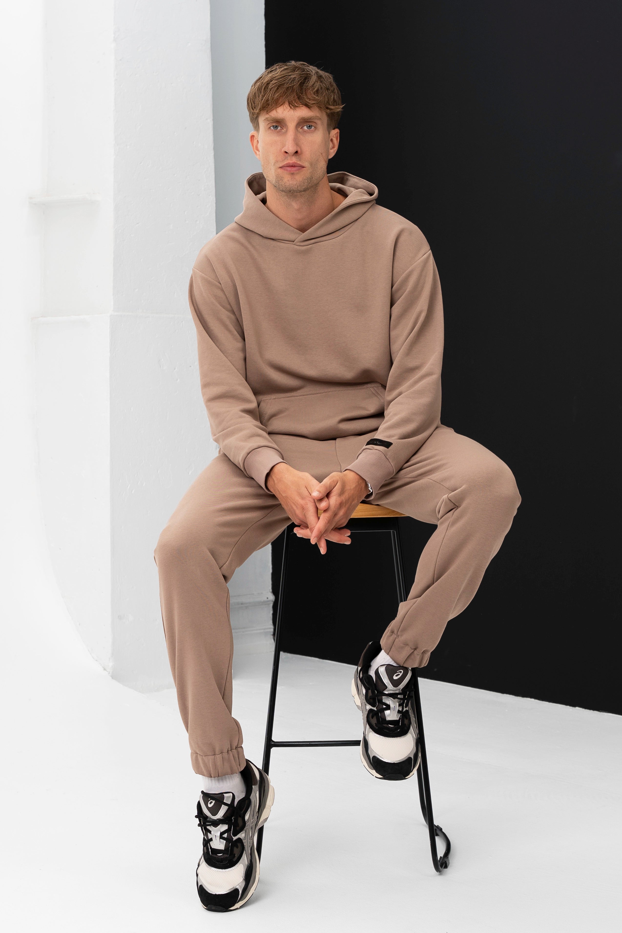 Modelis sėdi ant kėdės, dėvėdamas smėlio spalvos vyriškus treningus, rankos suglaustos ant kelių
