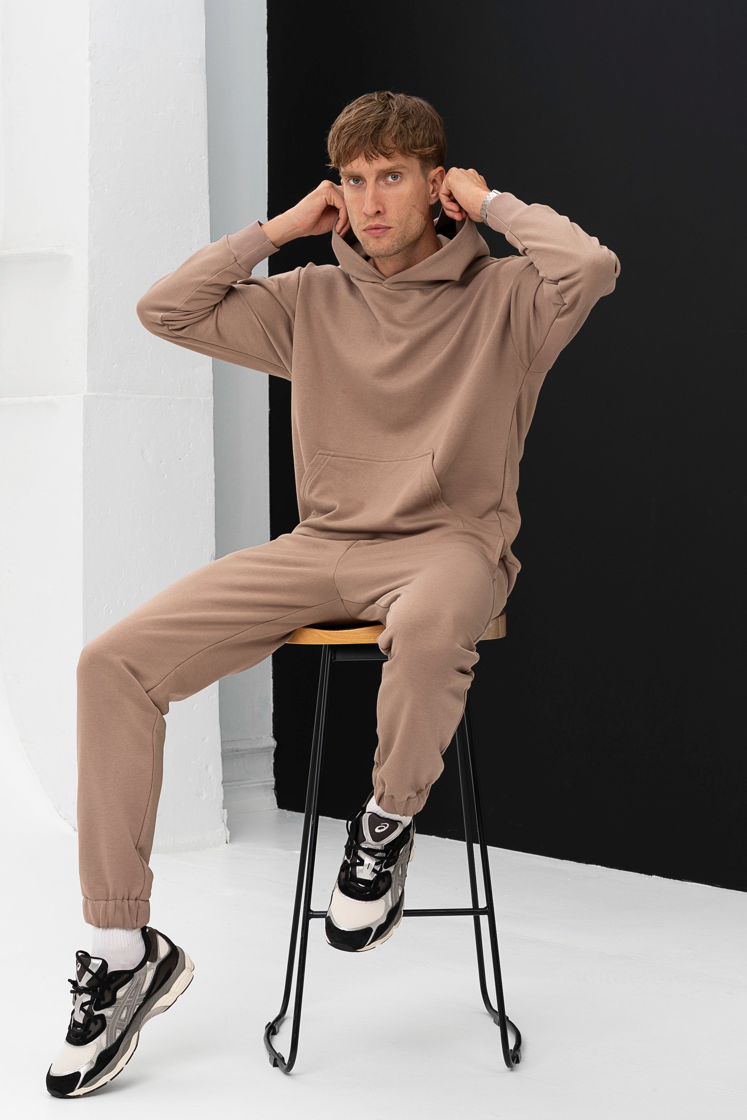 Modelis sėdi ant aukštos kėdės, dėvi patogius vyriškus treningus, rankomis prisilaiko džemperio kapišoną