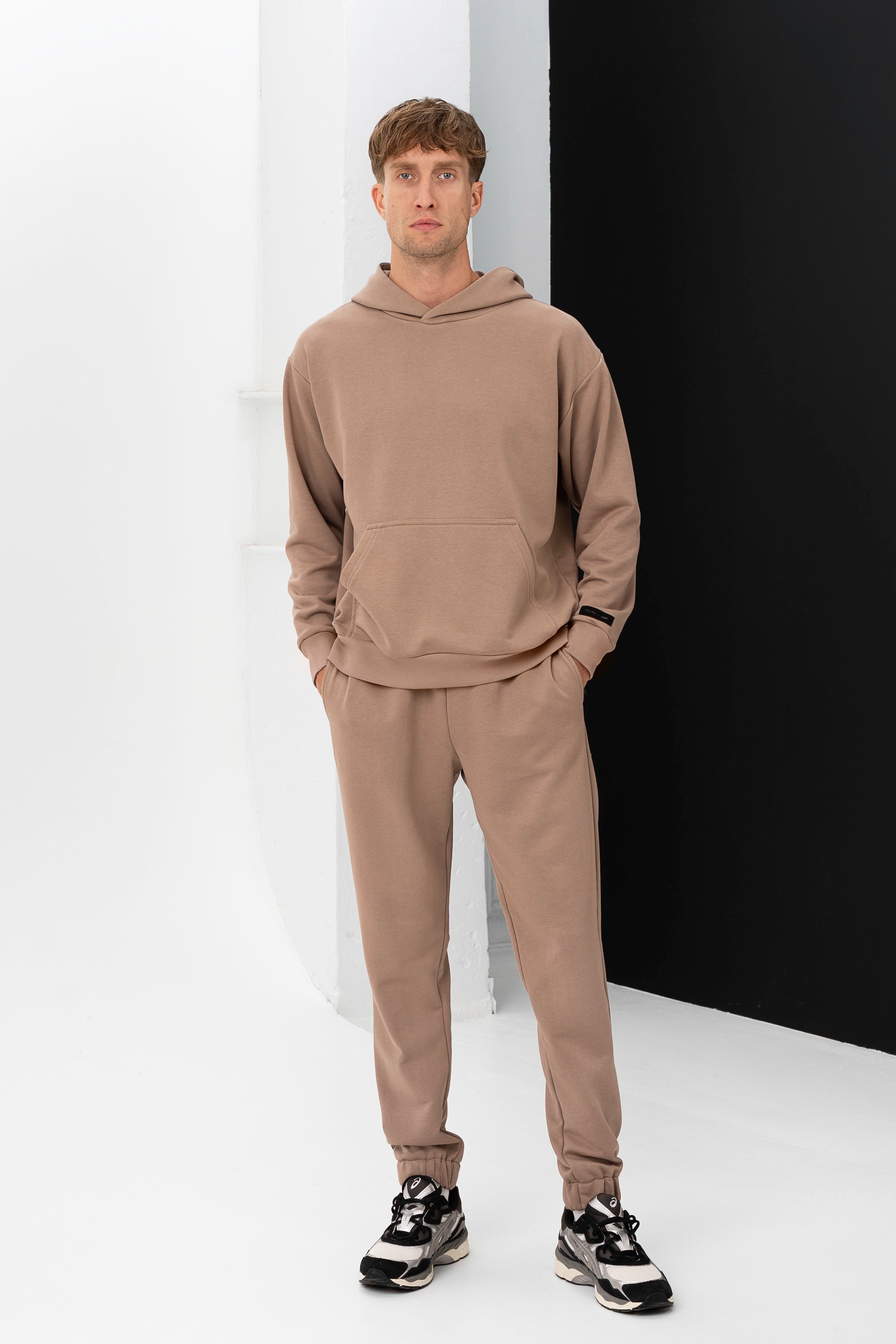Vyriški treningai – modelis stovi studijoje dėvėdamas smėlio spalvos džemperį su gobtuvu ir derančias kelnes