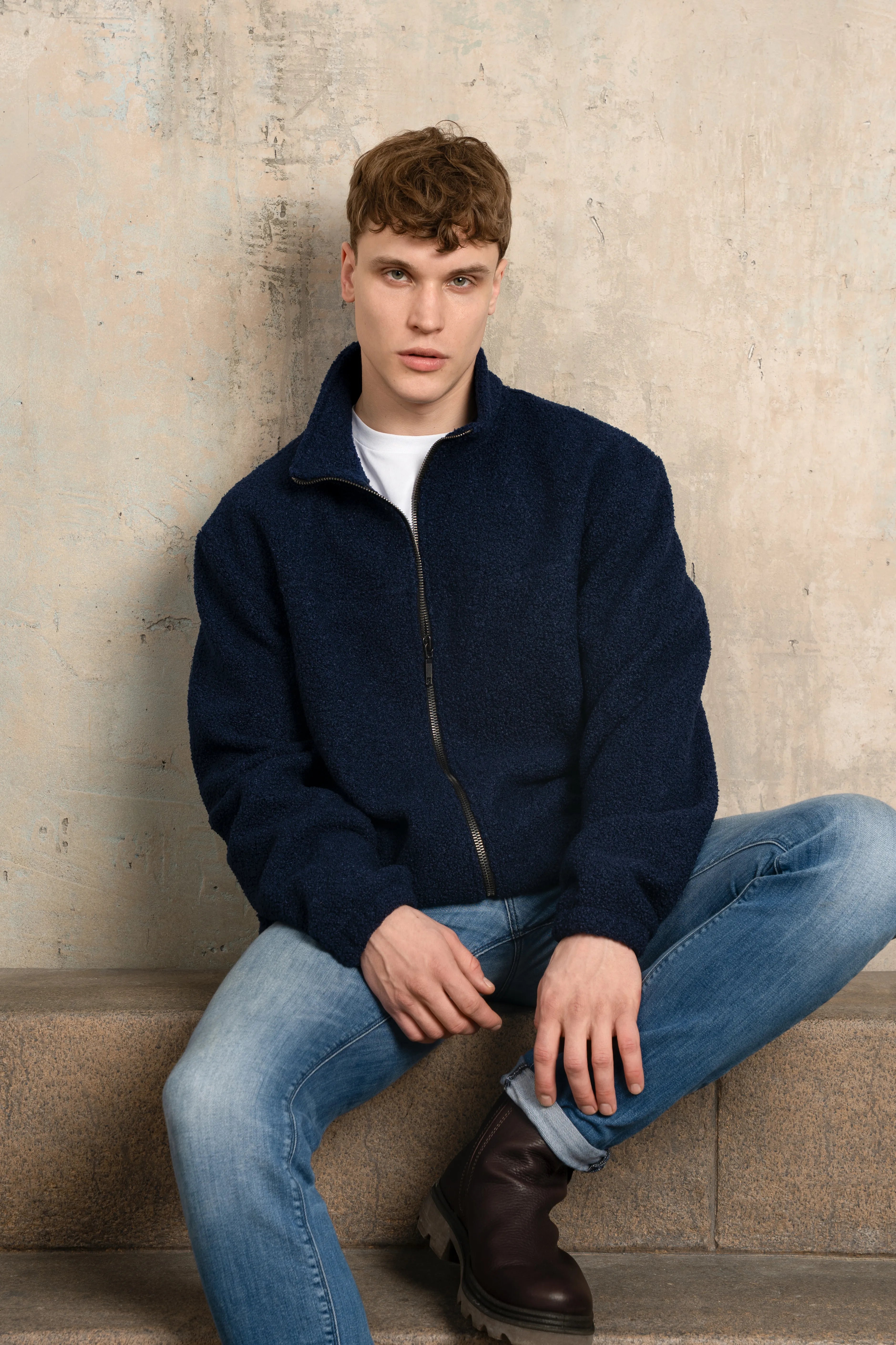 Tamsiai mėlynos spalvos vyriškas džemperis su pilno ilgio užtrauktuku, stačia apykakle, ilgomis rankovėmis ir laisvu siluetu, modelis nufotografuotas sėdintis