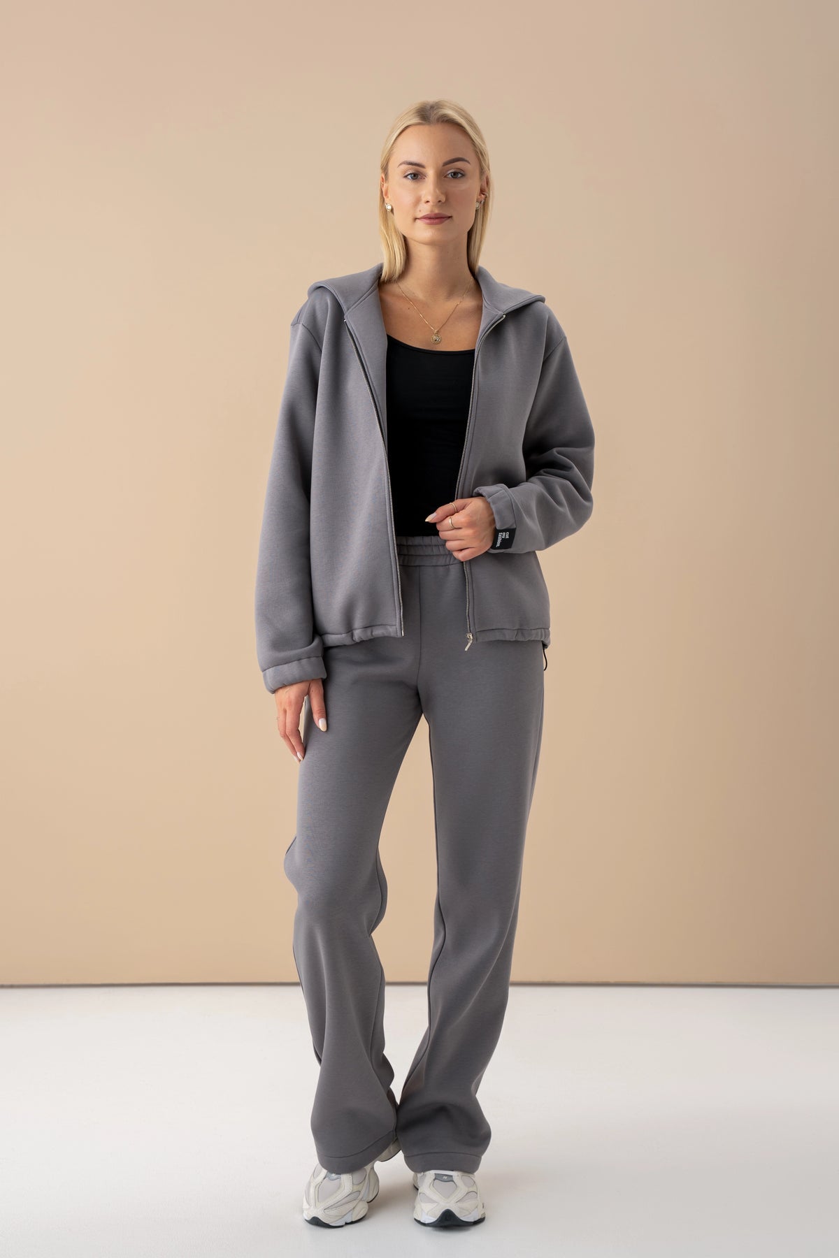 the woman is wearing grey cutmyfashion tracksuit with a black top, the zip-up sweatshirt is open, studio setting