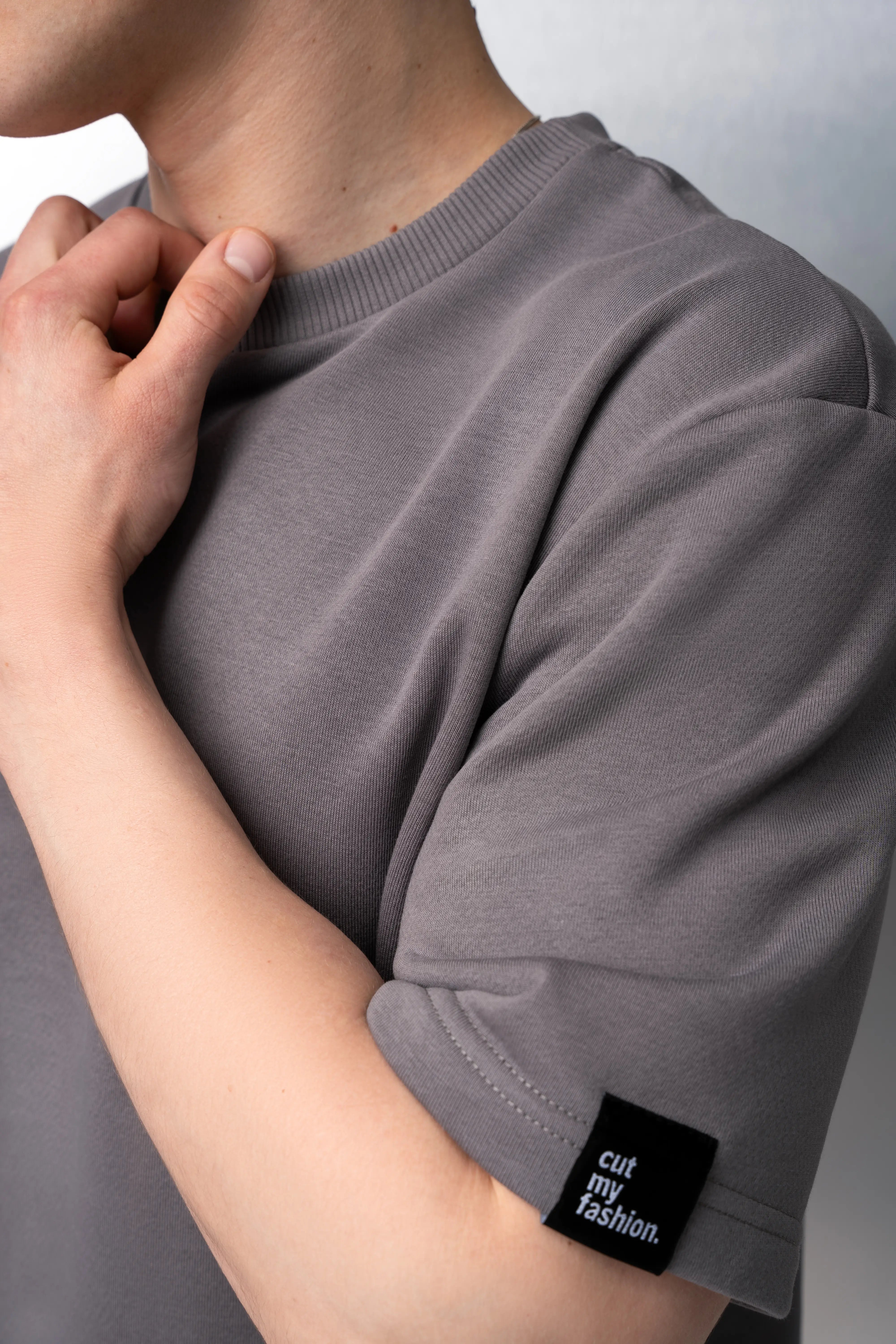 Pilki oversized marškinėliai vyrui iš be pūkelio audinio, su trumpomis rankovėmis, apvalia kaklo iškirpte ir juodu „cutmyfashion“ logotipu ant rankovės.