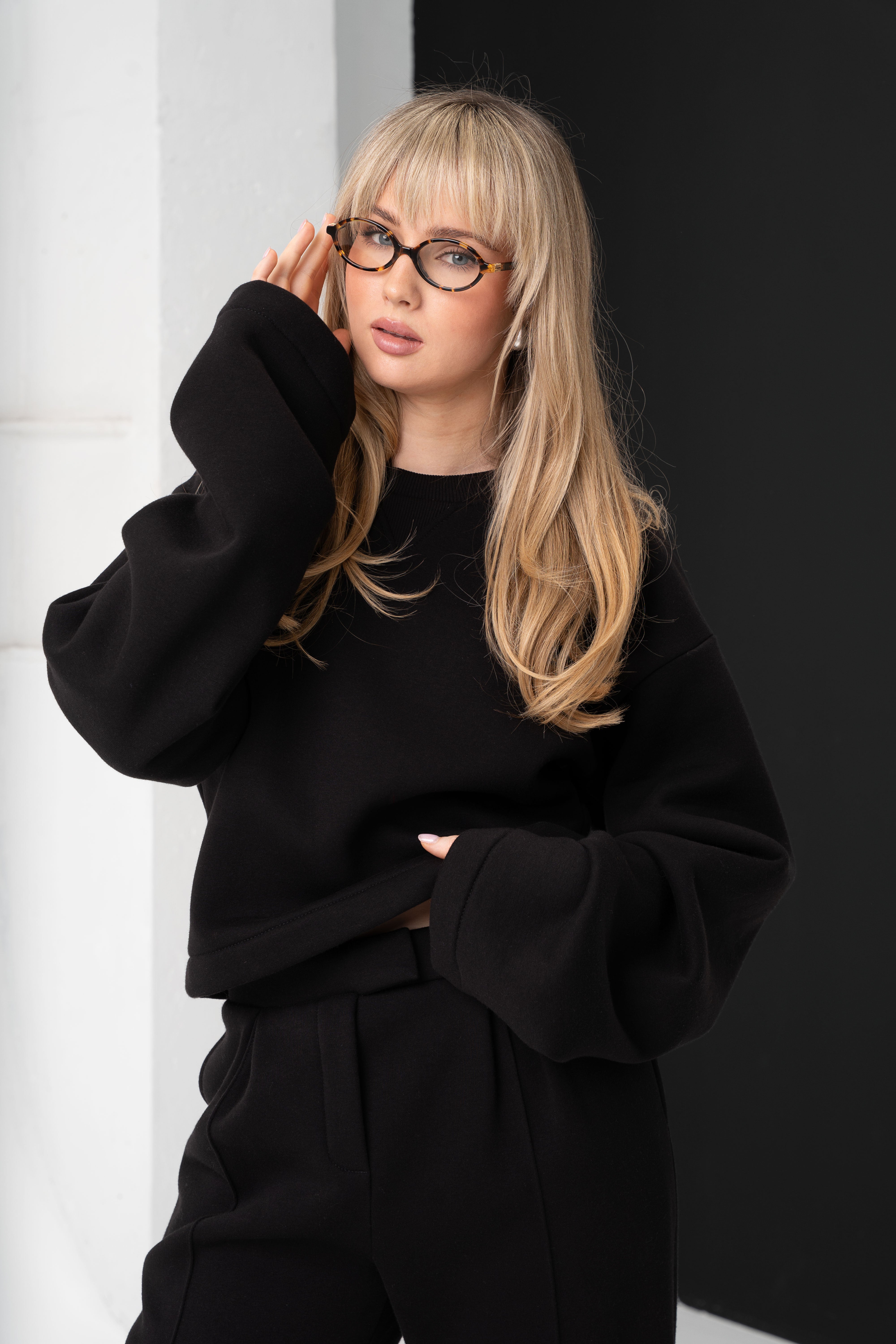 Stambiu planu modelis su akiniais liečia veidą, vilkėdama juodą „CMF by Kamilė“ džemperį