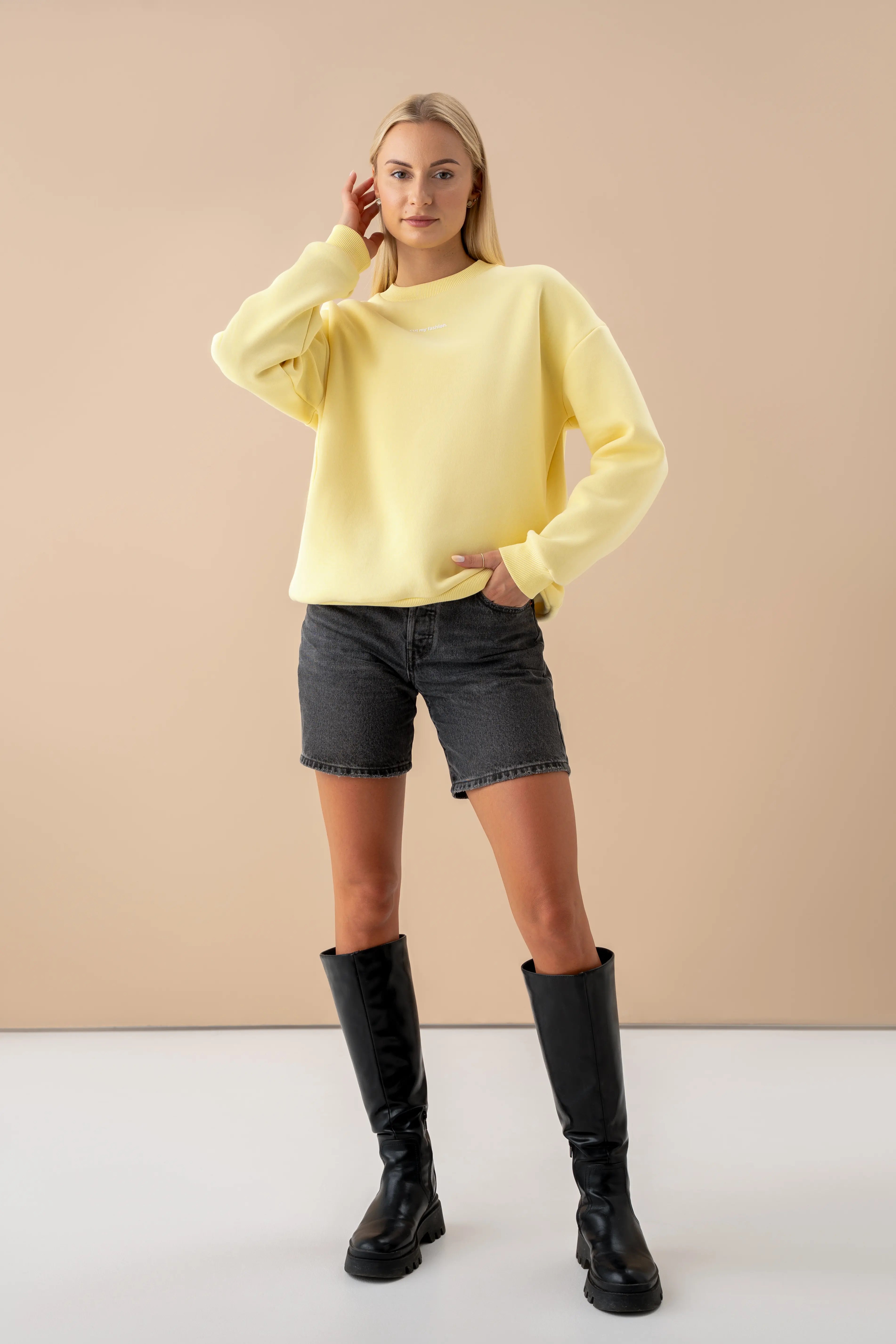 moteris su geltonu oversized cutmyfashion džemperiu iš OEKO TEX medvilnės stovi studijoje vilki džinsinius šortus ir juodus ilgaaulius batus pagaminta Lietuvoje