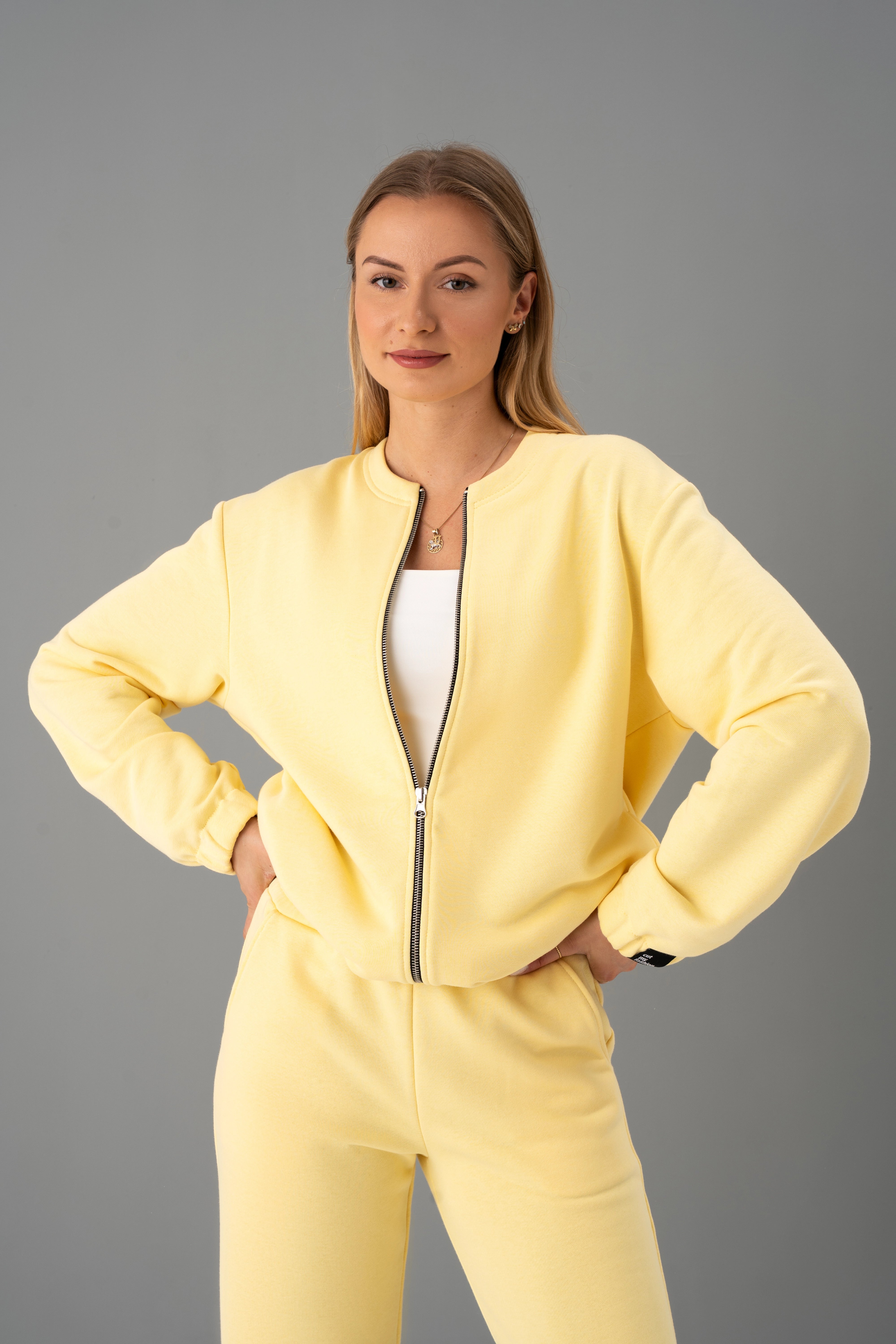 Modelis su geltonais cutmyfashion treningais Mėja CMF547-19 pagamintais Lietuvoje stovi studijoje, rankos ant klubų, žiūri į kamerą