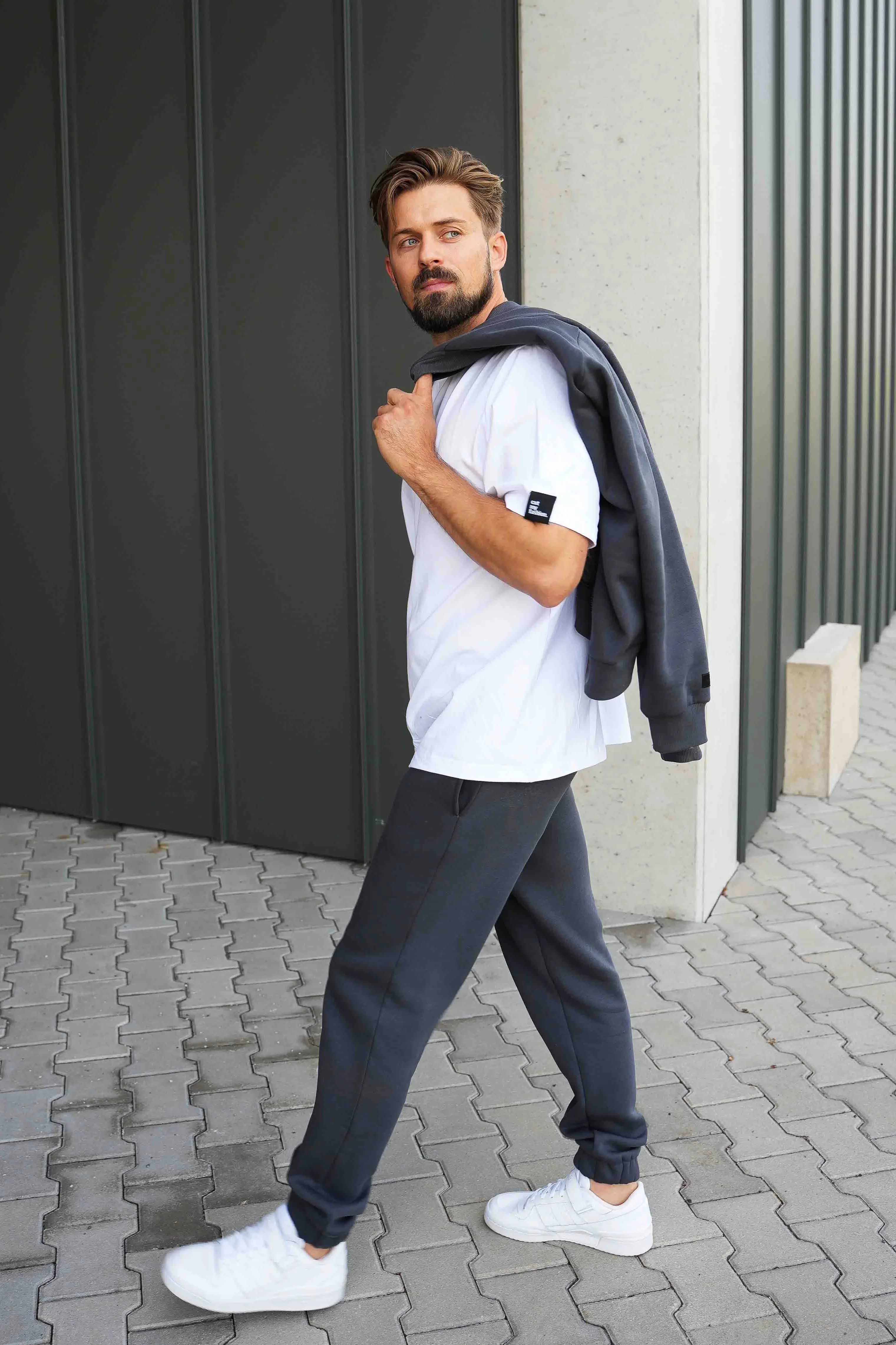 vyras vilki baltus cutmyfashion marškinėlius ir tamsias treningines kelnes, nešasi džemperį bomber stiliaus ant peties, eina miesto taku