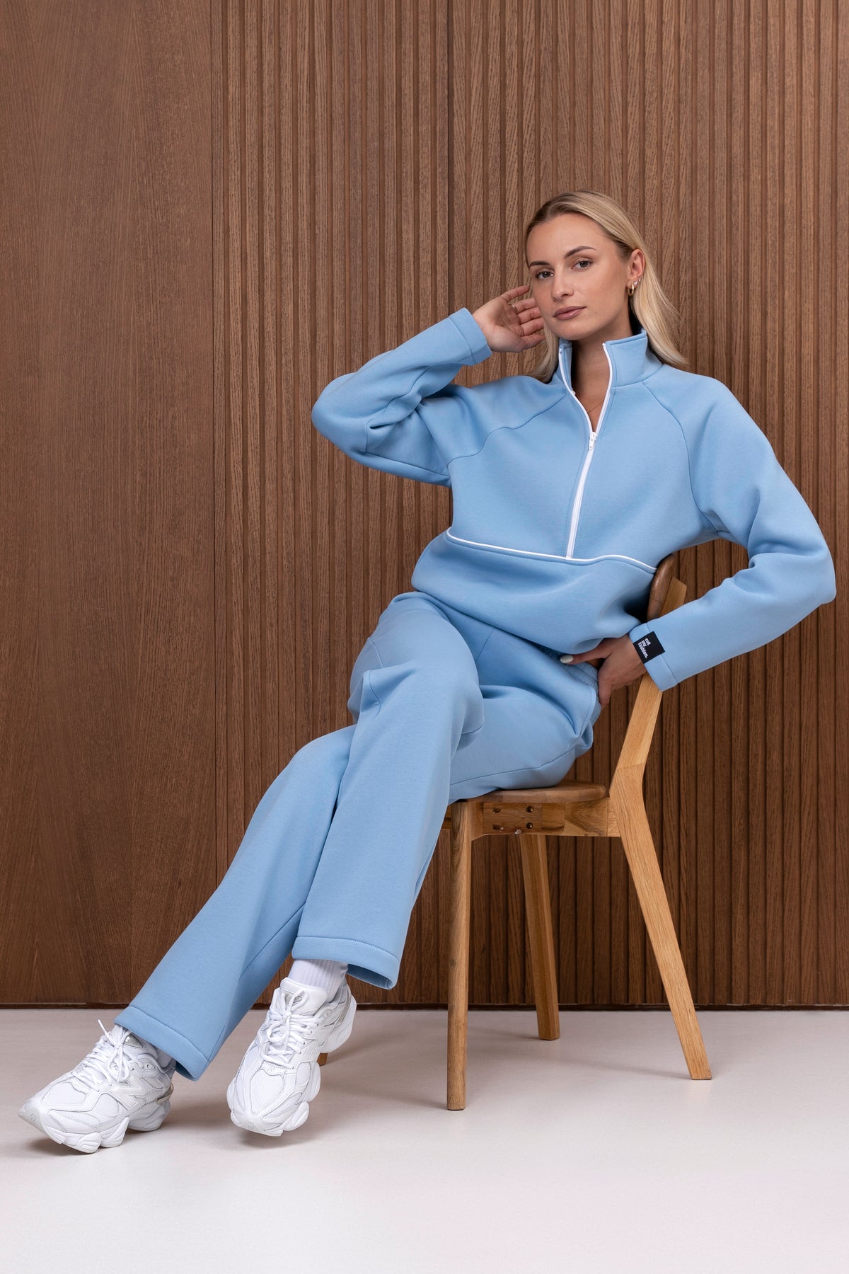Sėdinti moteris vilki dangaus žydrą laisvalaikio kostiumą su half zip džemperiu ir tiesiomis kelnėmis.