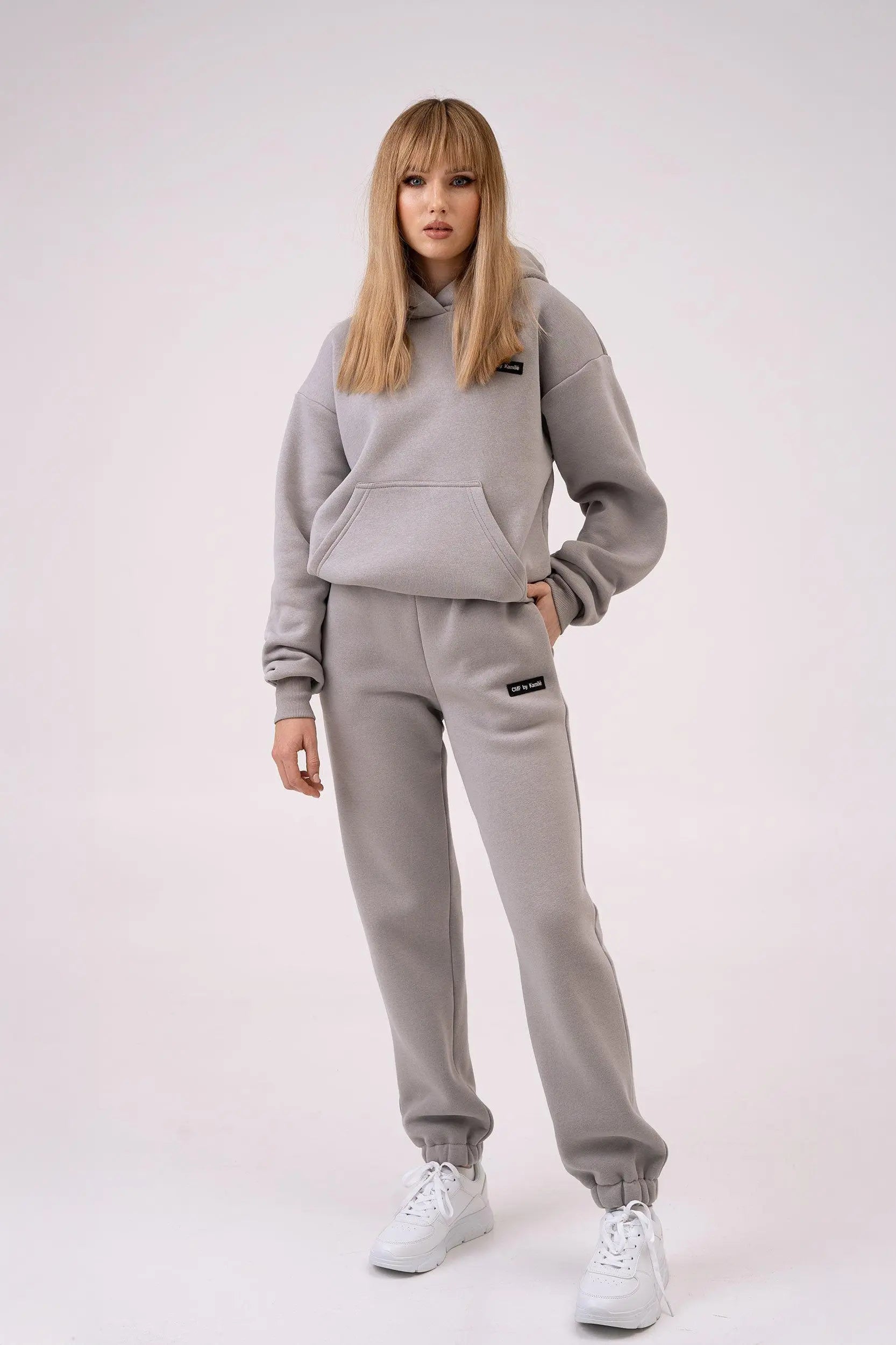 moteris dėvi cutmyfashion laisvalaikio treningus, stovi baltame fone, viena ranka kišenėje, kita nuleista.