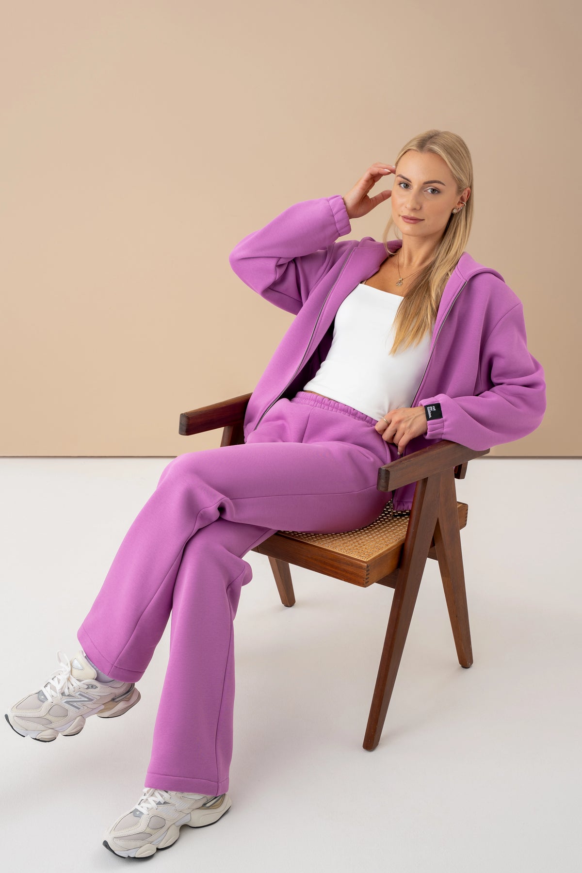 A woman is wearing violet tracksuits in the shade of cold beet soup with a white top, sitting on a wooden chair and looking at the camera